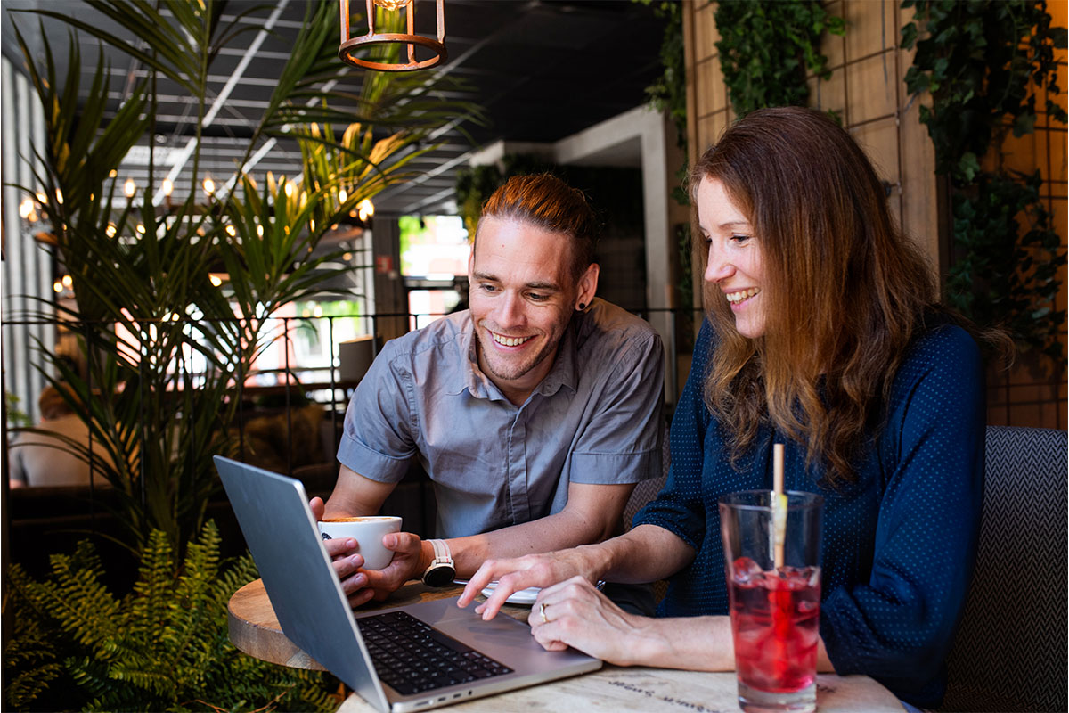 Two people sitting in a café, smiling while looking at a laptop together.