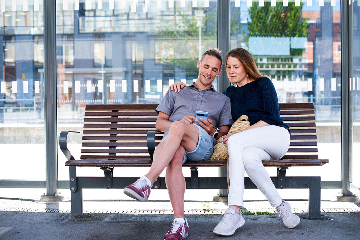 Two people sitting on a bench at a bus stop, looking at a smartphone together.
