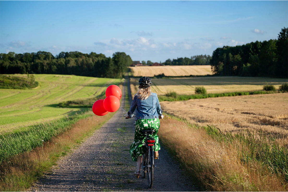Person cycling down a rural path, holding three red balloons, with fields and trees around.